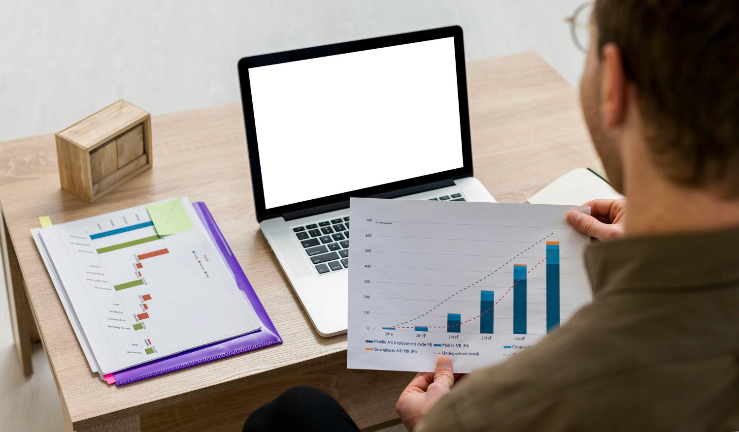  A man is holding a chart while sitting at a desk with a laptop in front of him, analyzing data and making decisions.