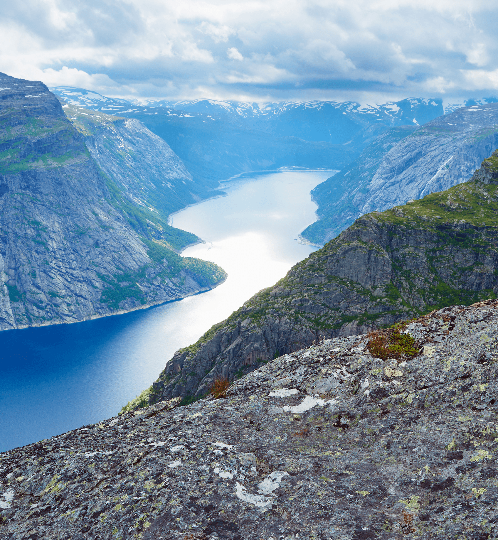 Mountain fjord with steep rocky cliffs and a winding blue lake under a cloudy sky.