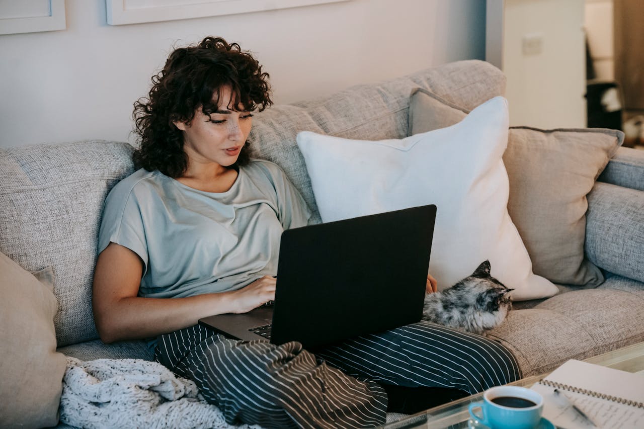 Picture of a woman sitting on the couch with a laptop on her lapt and a cat next to her