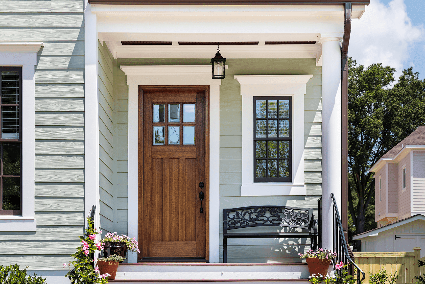 A charming front entrance featuring a wooden door with glass panes, surrounded by sage-green siding and white trim. A black decorative bench sits on the porch alongside potted flowers, creating a welcoming atmosphere.