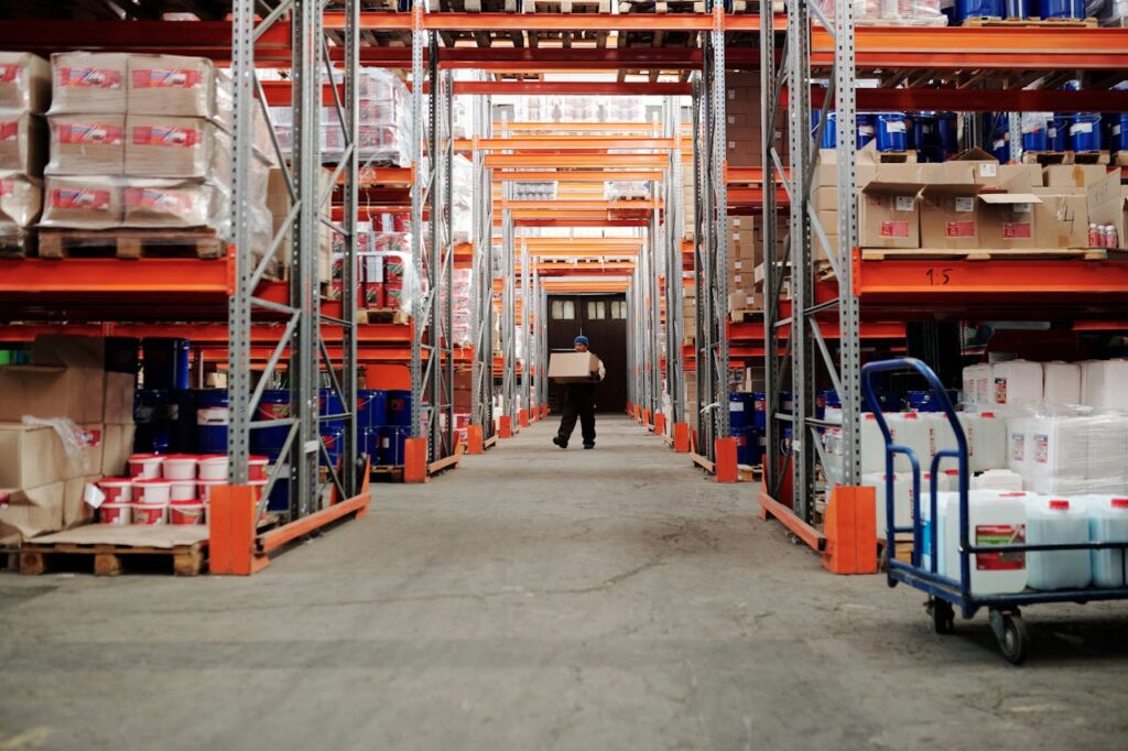 Long view down a well-stocked warehouse aisle with tall industrial shelving units, filled with various boxes and containers. A person carries a box in the distance, conveying efficient logistics and inventory management