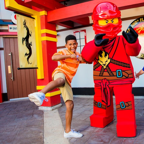 A person poses playfully with a large, red ninja LEGO character outside a themed building decorated with a dragon banner.
