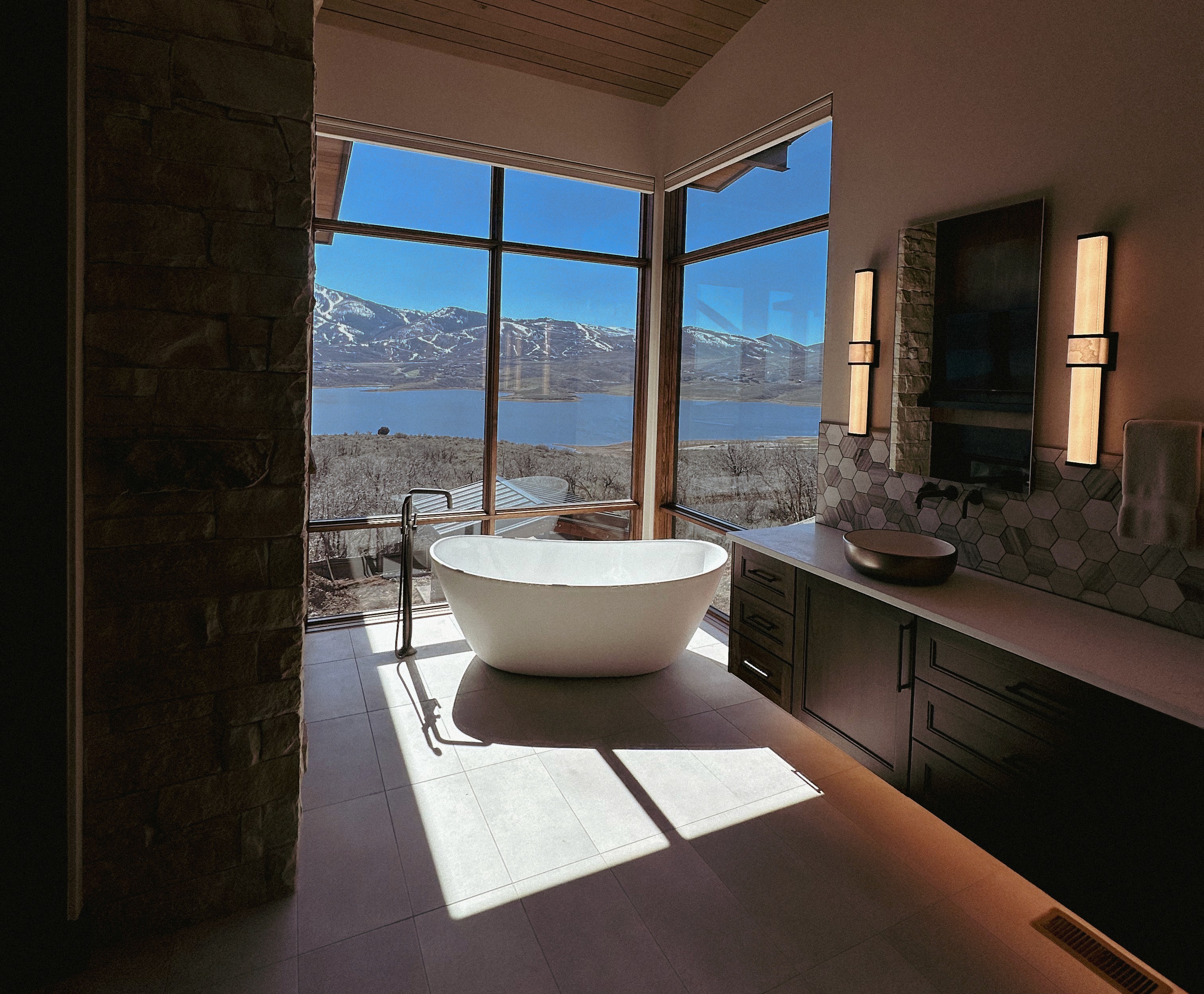 A spacious industrial-style interior with exposed brick walls, large windows, and natural light pouring in during sunset. Wooden desks with plants and workspace materials are arranged along the walls, with a view of greenery and an outdoor seating area.