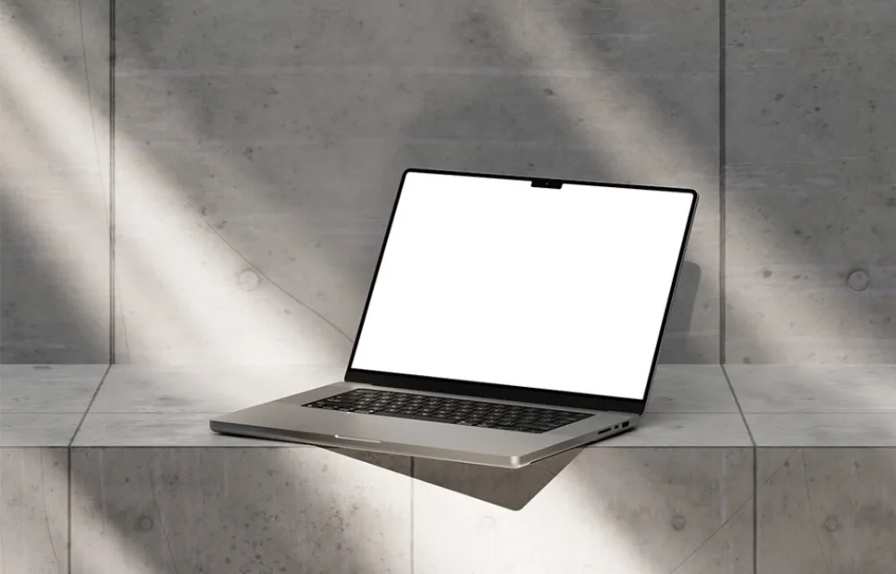 MacBook Pro mockup with a blank white screen, displayed on a concrete surface. The background features a textured concrete wall with soft sunlight casting natural shadows.