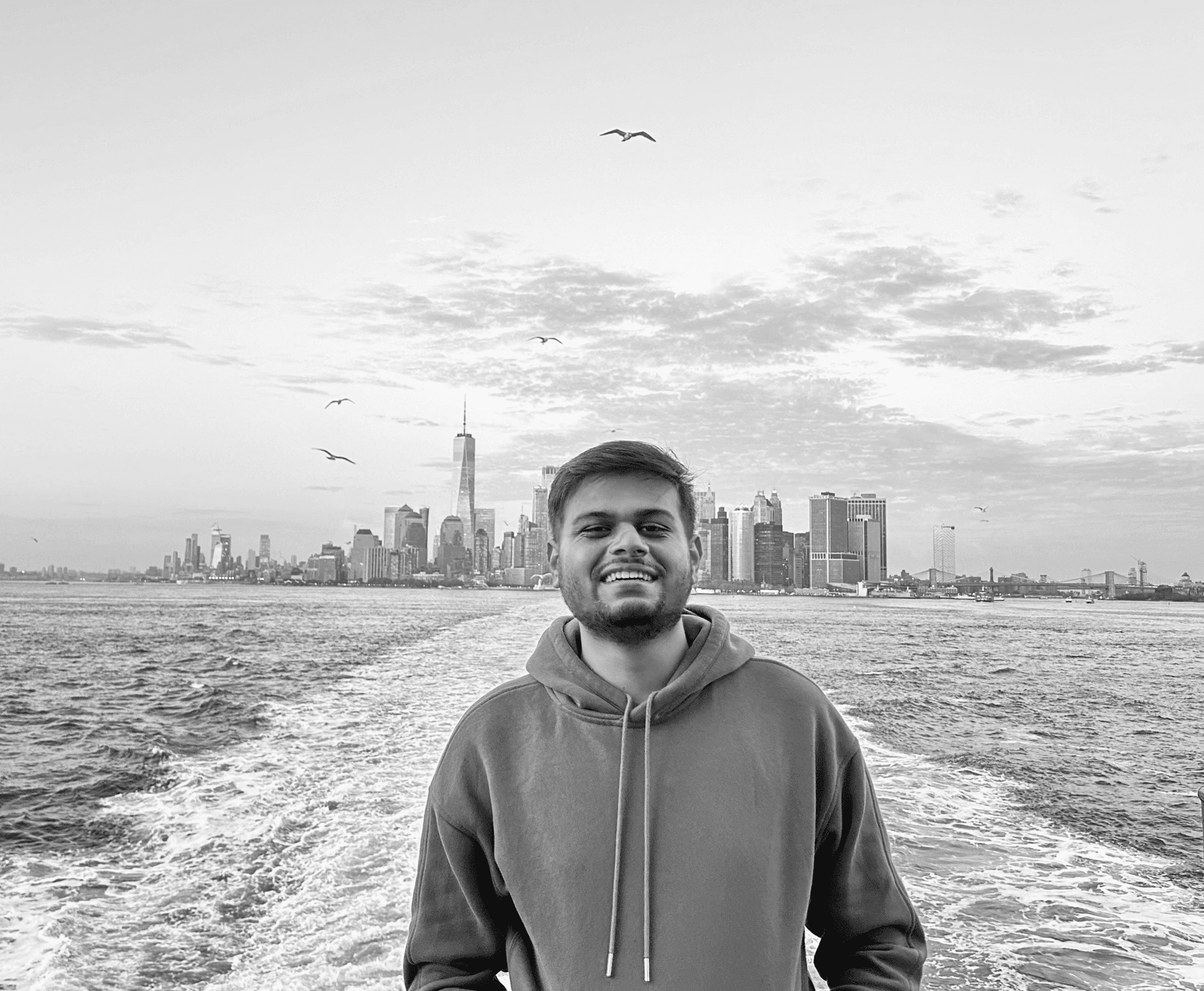 Amish in a hoodie stands smiling on a boat with a Ney York city skyline and birds in the background.
