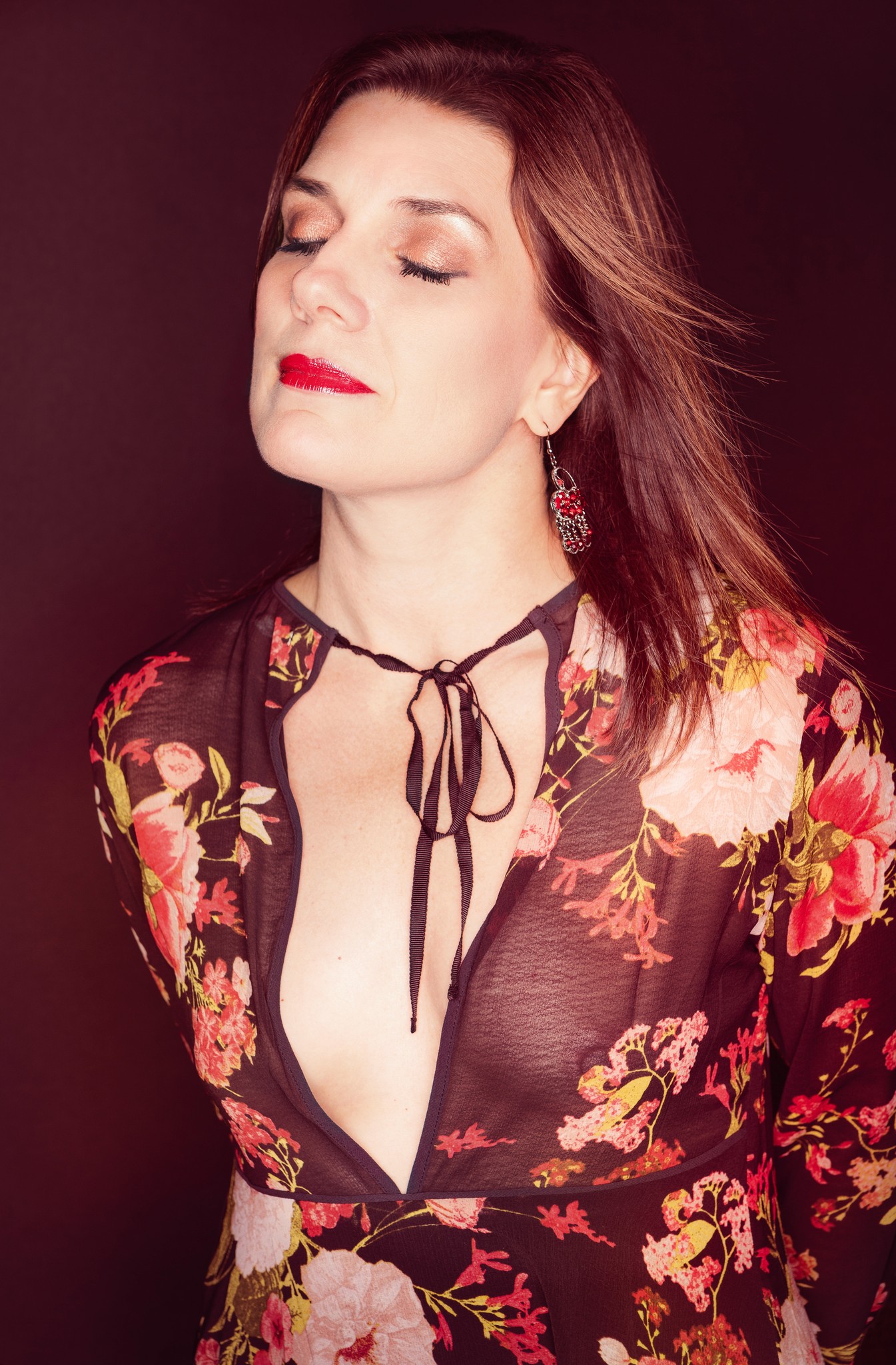 Editorial portrait of a woman in a floral dress with eyes closed, photographed against a dark background with soft, directional light.
