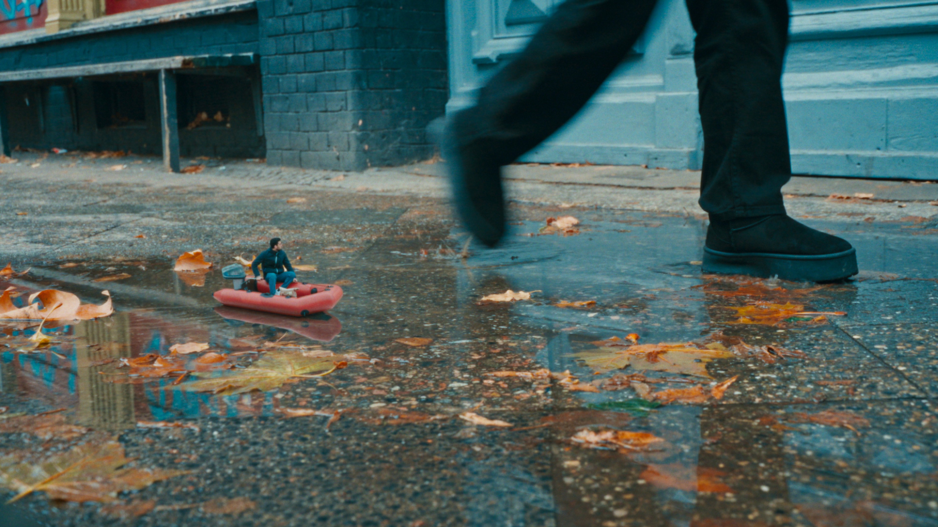 A miniature protagonist crosses a puddle in a tiny boat while a life‑size passerby walks past — a metaphor for precarious journeys and vulnerability.