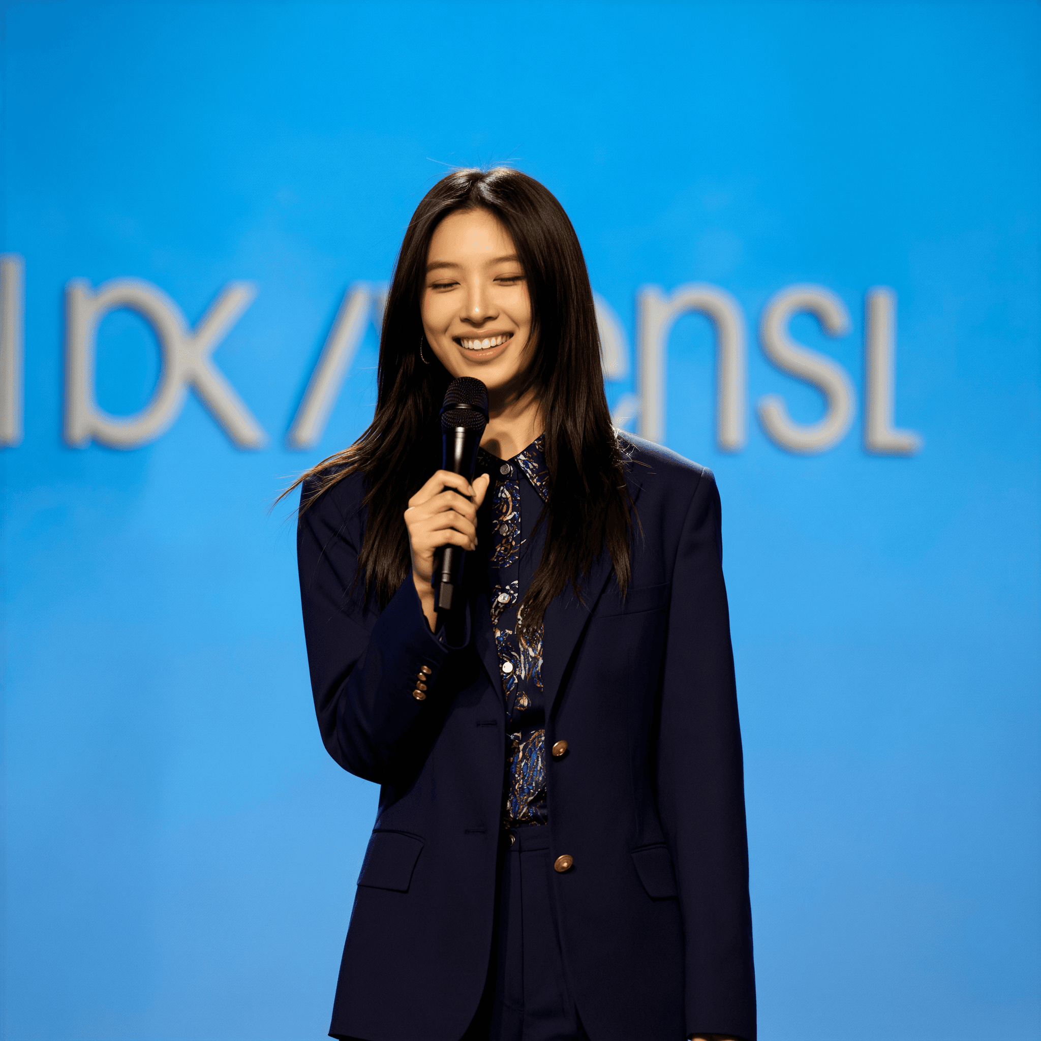 KI-generiertes weibliches Model spricht lächelnd in ein Mikrofon auf einer Bühne; trägt einen dunkelblauen Business-Anzug vor blauem Präsentationshintergrund – Szene einer Konferenz, Moderation oder Keynote.