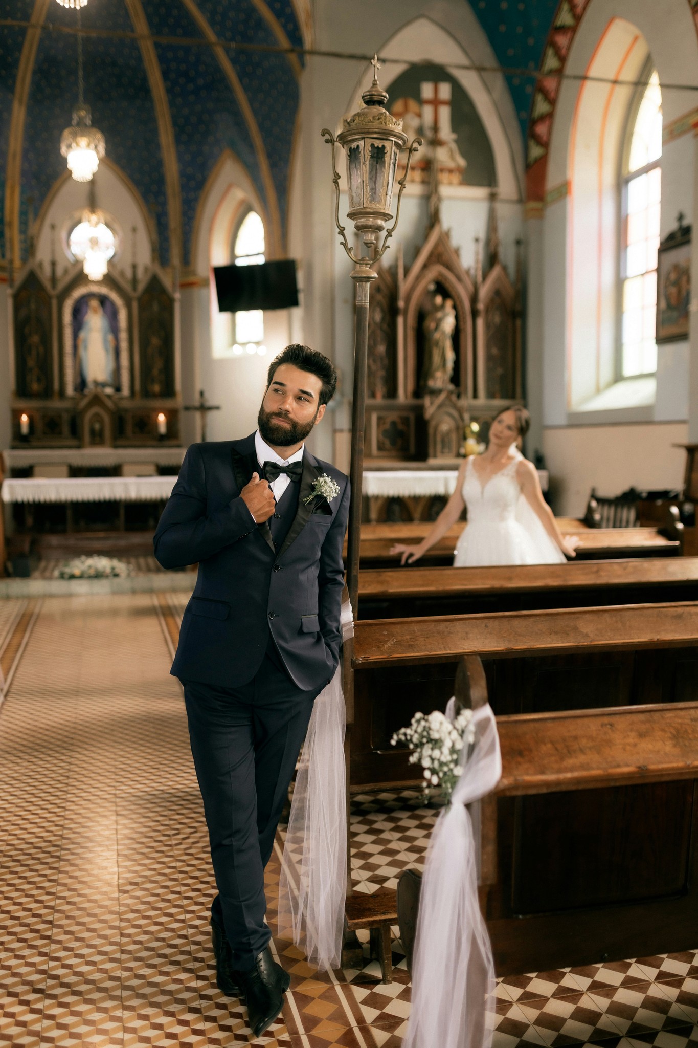Groom adjusting his jacket in a historic church aisle with the bride seated among wooden pews in the background
