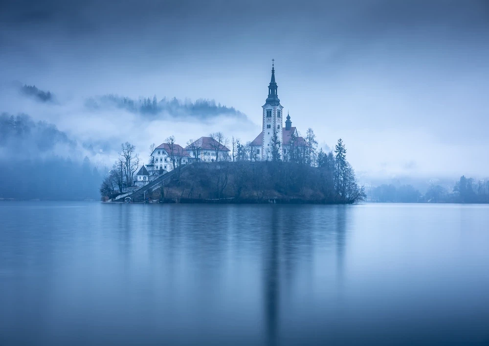 Lake Bled Island, Slovenia, surrounded by misty turquoise waters and lush green trees on a rainy, atmospheric day.