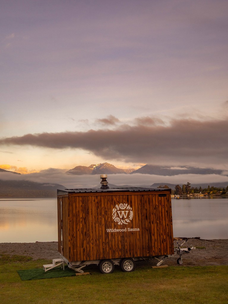 A wooden sauna trailer sits by a tranquil lake at sunset, with misty mountains in the background. The sky is pastel with scattered clouds. Serene and peaceful.