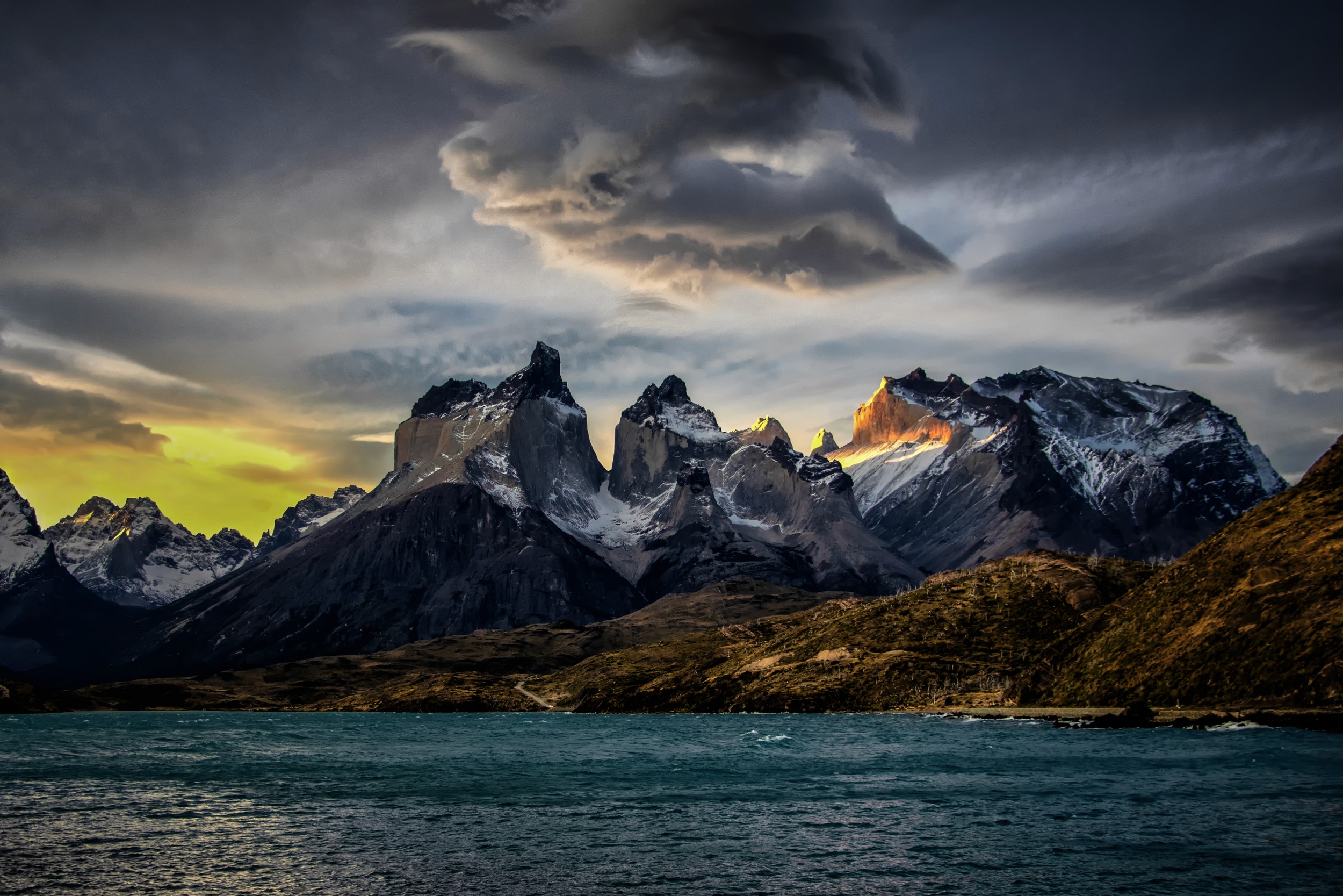 The Torres del Paine range under a dramatic evening sky, with dark clouds and deep turquoise water in the foreground