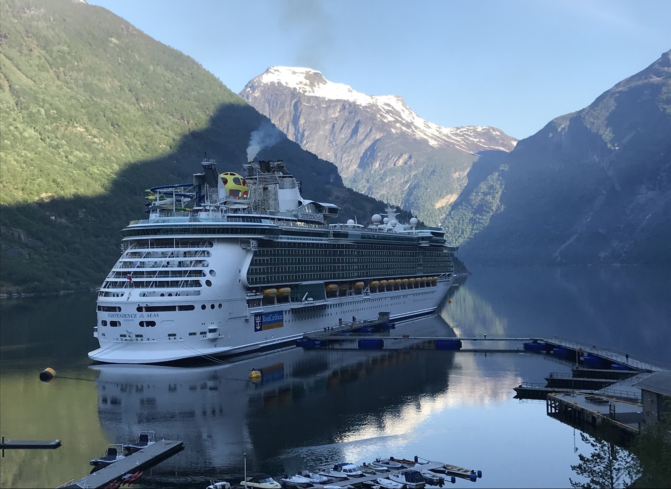 Cruise ship docked at a floating pier in Geiranger fjord, surrounded by steep mountains and calm water reflecting the vessel and landscape.