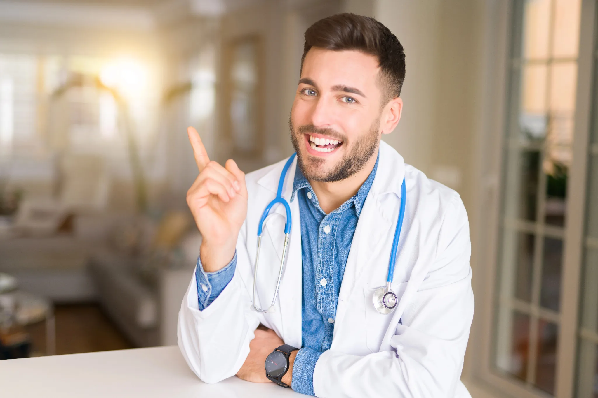 A smiling male doctor wearing a white lab coat and blue stethoscope gestures enthusiastically while seated at a desk in a clinical setting.