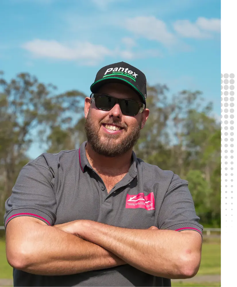 Liam Garner, Director and Nominee Supervisor of Queensland Quality Roofing, standing outdoors in a grey company polo shirt with arms crossed, smiling confidently under a clear blue sky. A trusted expert in roofing services across Brisbane & Southeast Queensland.
