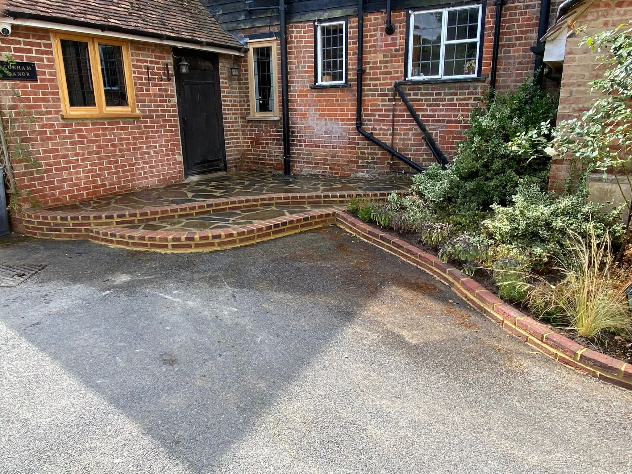 A brick pathway leads to a doorway, surrounded by a garden and part of a house, with sunlight casting shadows.