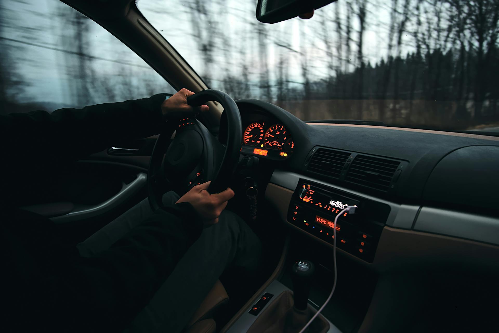  A low-light image showing the dashboard and interior of a car while driving. The driver's hands are on the steering wheel, and the speedometer and radio display are illuminated with orange backlighting. The background visible through the windshield shows blurred trees, indicating motion. A charging cable is plugged into the console.
