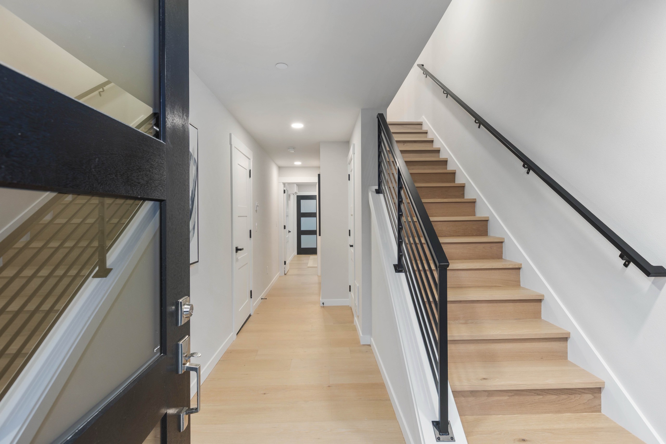 Modern luxury home interior hallway with light oak hardwood floors, black metal stair railing, recessed lighting, and clean white walls leading to multiple rooms