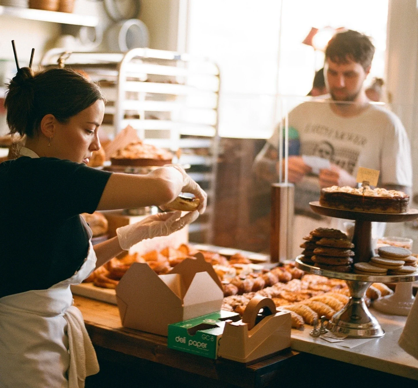 Bakery worker in gloves packaging pastries while colleague works in background; "deli paper" box visible on counter.