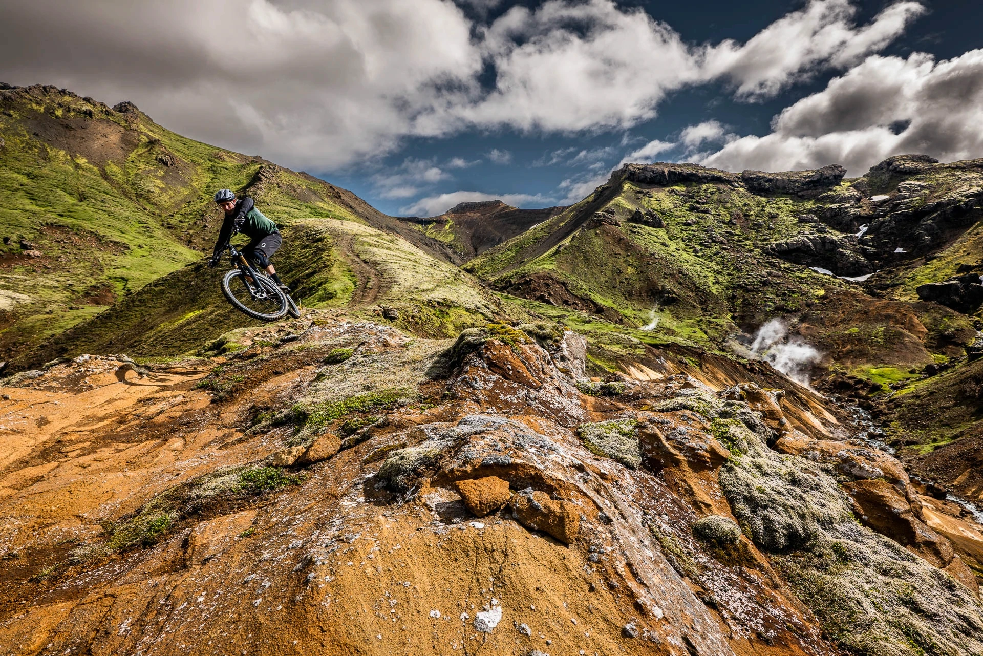 Mountain biker descending a rocky trail above geothermal steam and green hills.