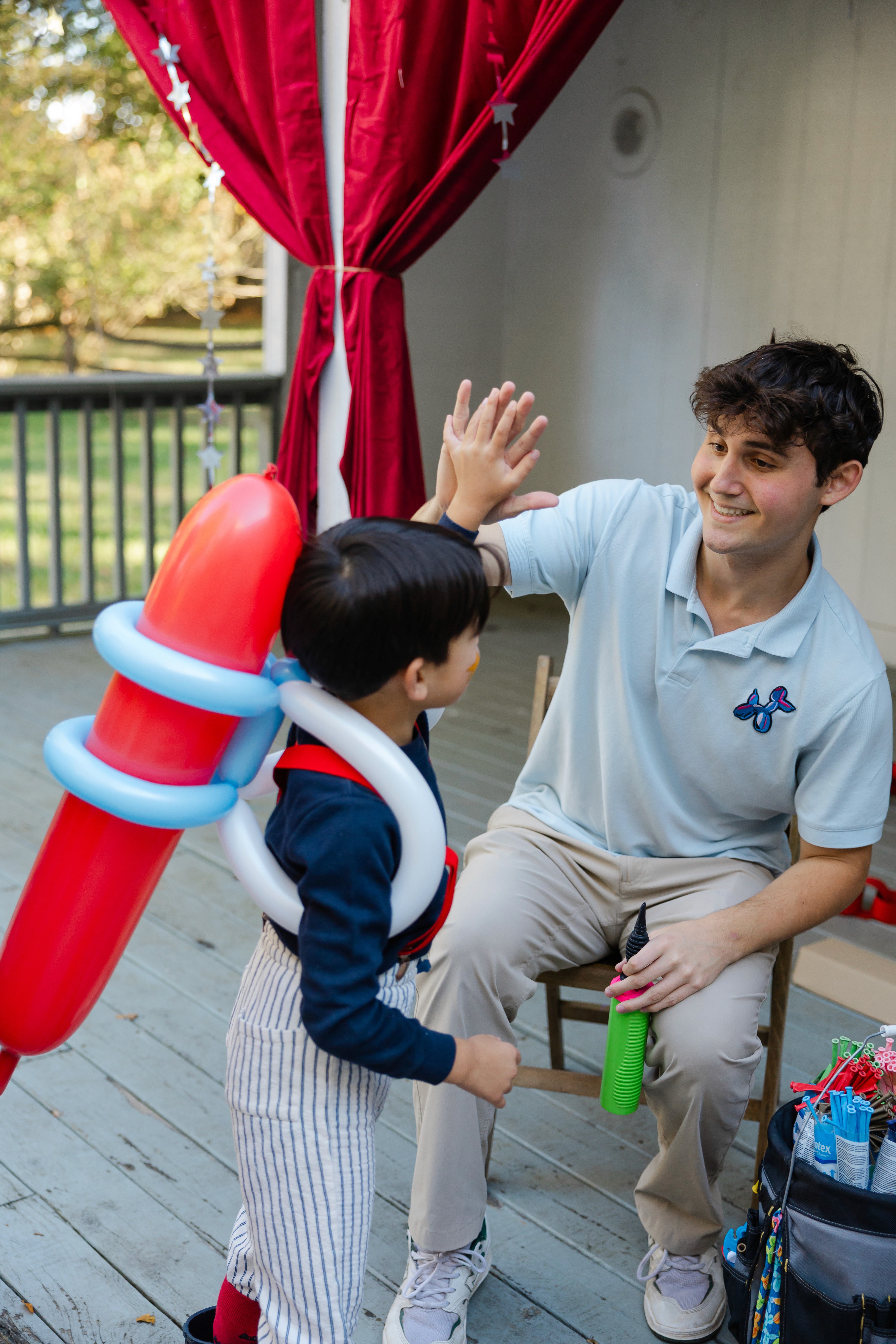 A young boy wearing a balloon rocket backpack gives a high-five to Daniel in a light blue polo shirt, seated on a porch with red curtains in the background, enjoying a playful moment outdoors