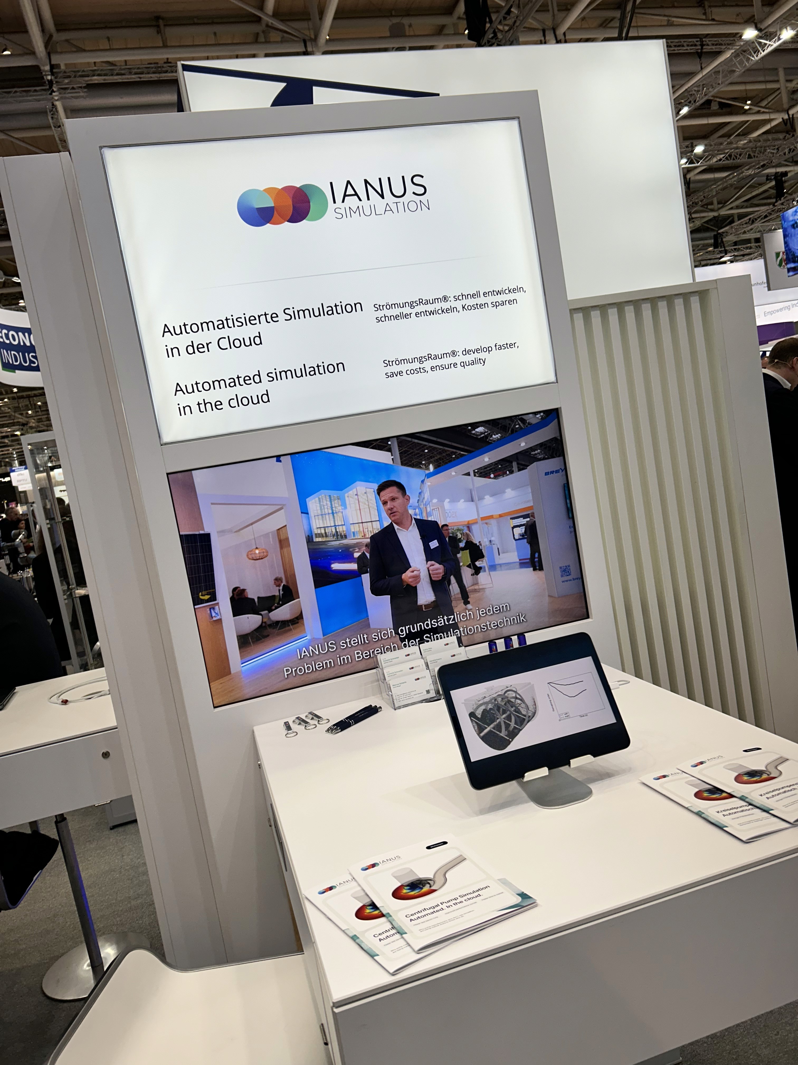 Mann im Anzug präsentiert auf einem Messestand vor einem großen Bildschirm die Lösungen von IANUS Simulation