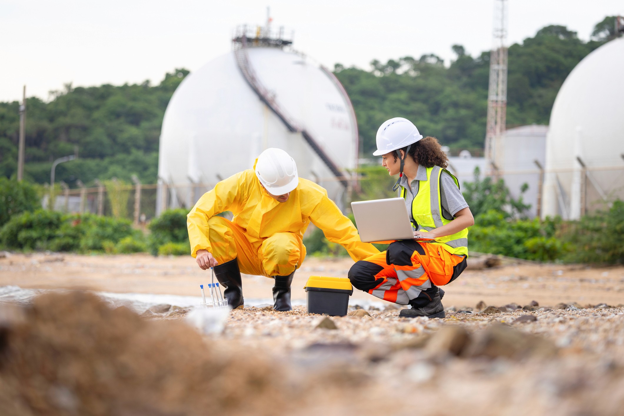 Workers in protective gear examine the ground near large storage tanks on a cloudy day.