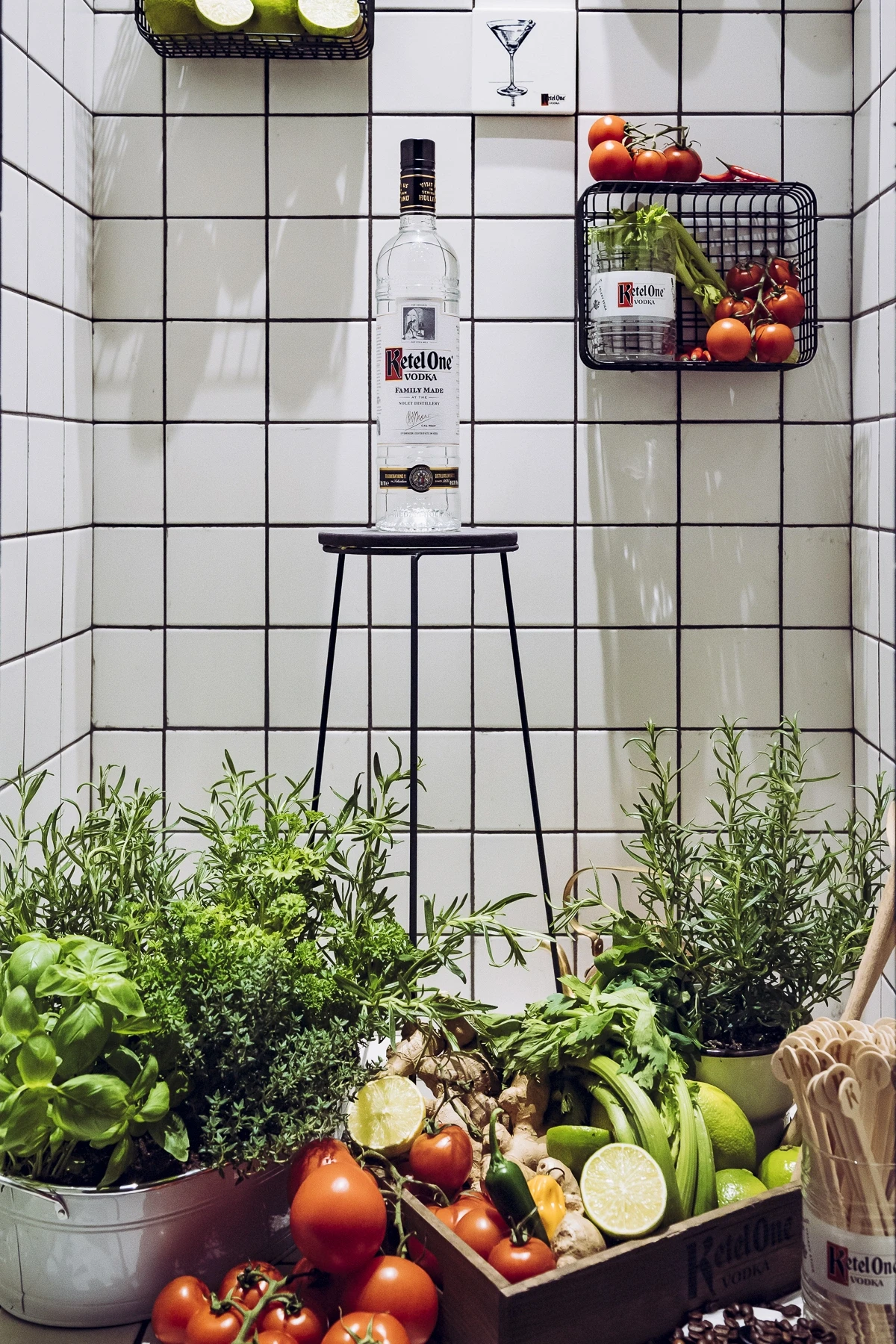 Ketel One vodka bottle on a black stool against white tiles at DIAGEO BCB19, surrounded by fresh herbs, tomatoes and lime baskets.