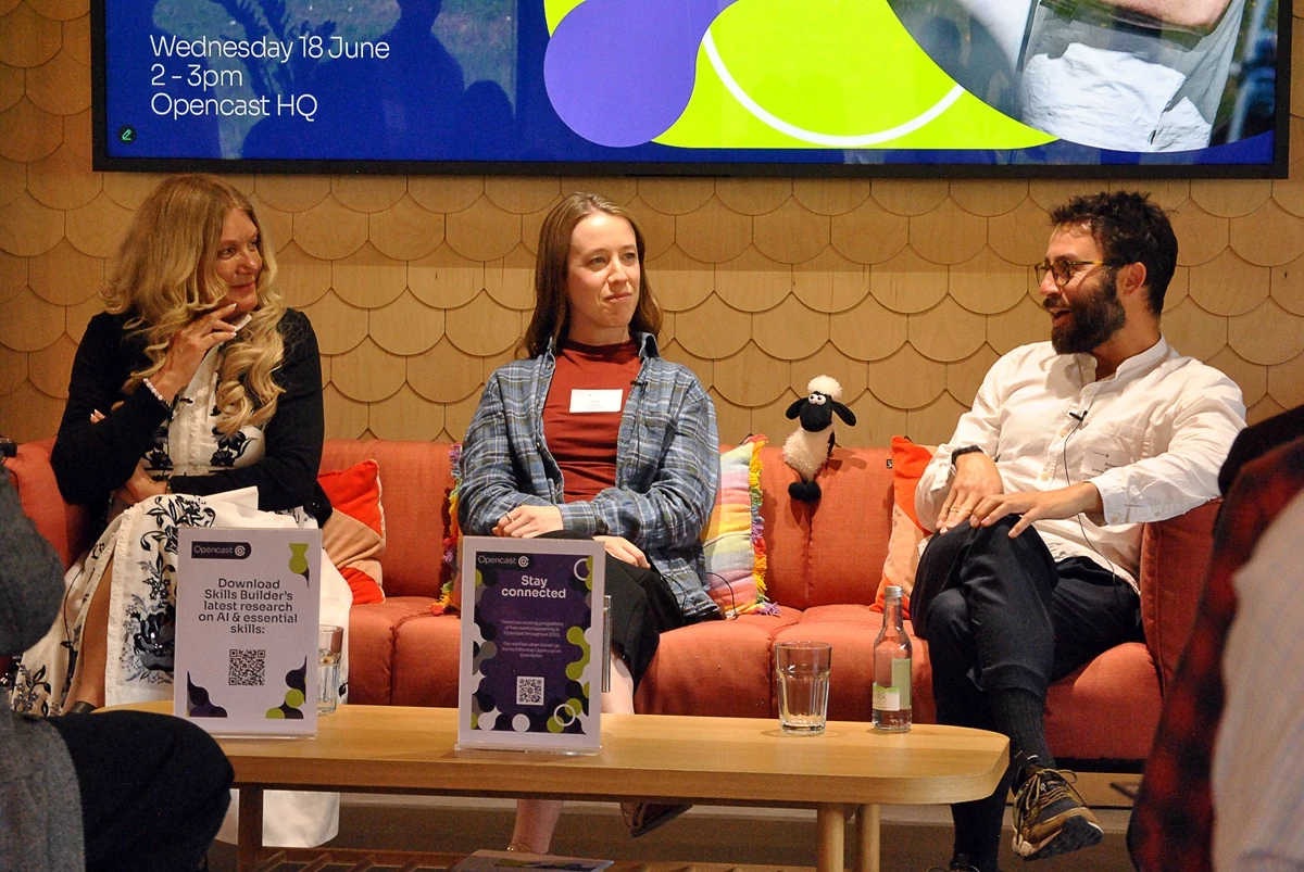 Three people seated on a sofa during a panel discussion, with a large screen behind showing event details and a coffee table in front with signs, glasses, and a bottle.