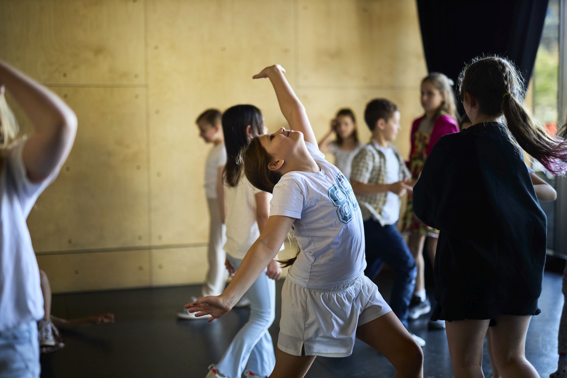 A group of children take part in a movement activity in a studio, with one child leaning back dramatically while others move around the space.