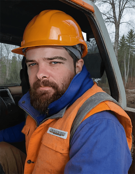 Algonquin Septics team member in orange safety gear inside an excavator, representing professional septic system services in forested Northern Ontario.