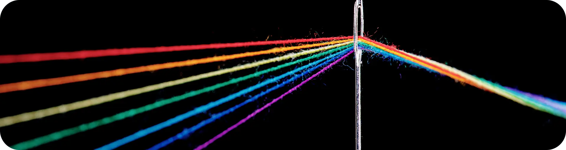 Fotografia em close com fundo preto. Seis fios de linha em diferentes cores (vermelho, laranja, amarelo, verde, azul e roxo) vêm separados do lado esquerdo, convergem e passam todos juntos pelo buraco de uma única agulha de costura prateada, saindo unidos do lado direito.