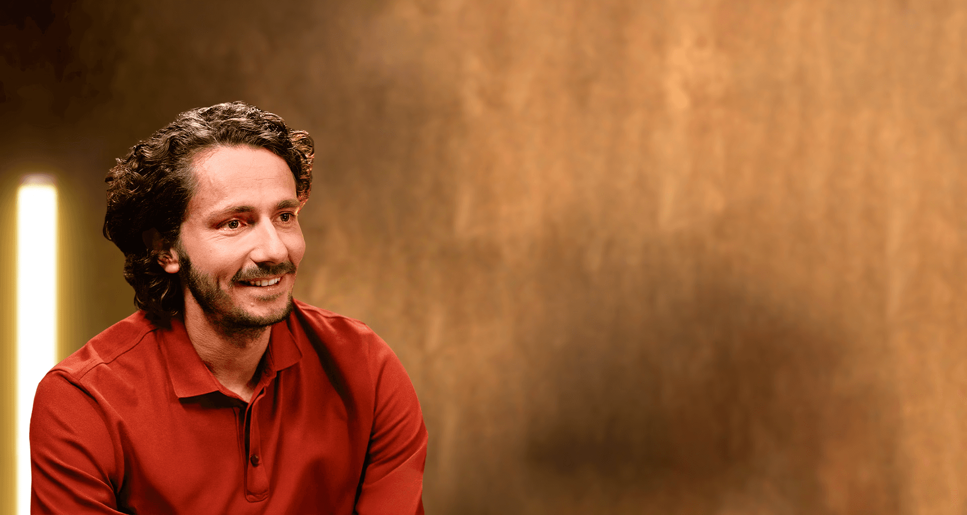 Portrait professionnel et souriant de Guillaume Pley, fondateur du média digital Legend, dans son studio d'enregistrement. Il est vêtu d'un polo rouge terre cuite. Le sujet regarde vers la droite, laissant un large espace négatif à droite de l'image. L'arrière-plan est un mur texturé, flou, dans des tons chauds (or, bronze, marron), éclairé par une colonne lumineuse verticale à l'extrême gauche, créant une ambiance de studio d'interview et de podcast haut de gamme. 