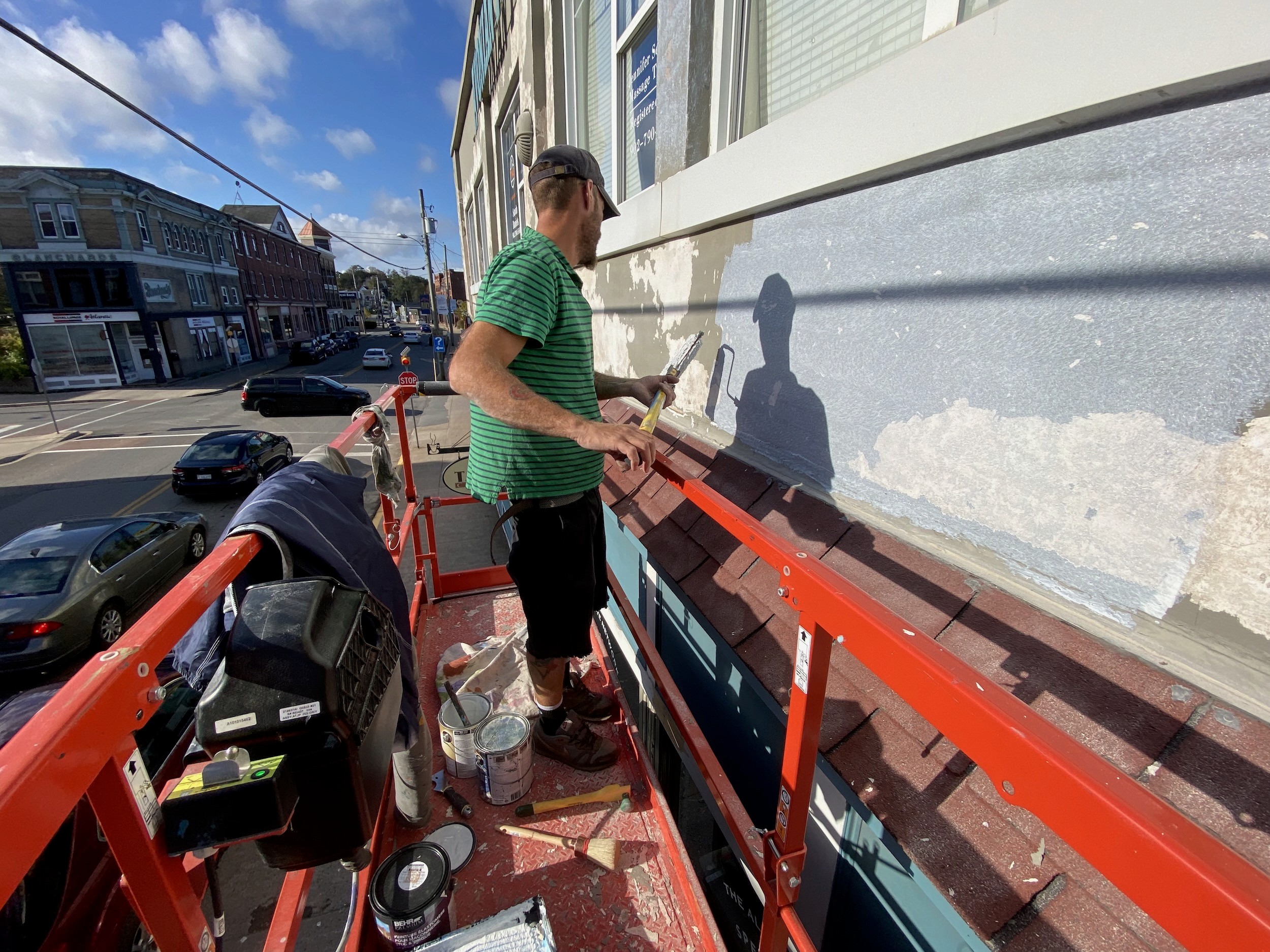 From a red lift, a man scrapes and paints the upper wall of a building above a small-town street.