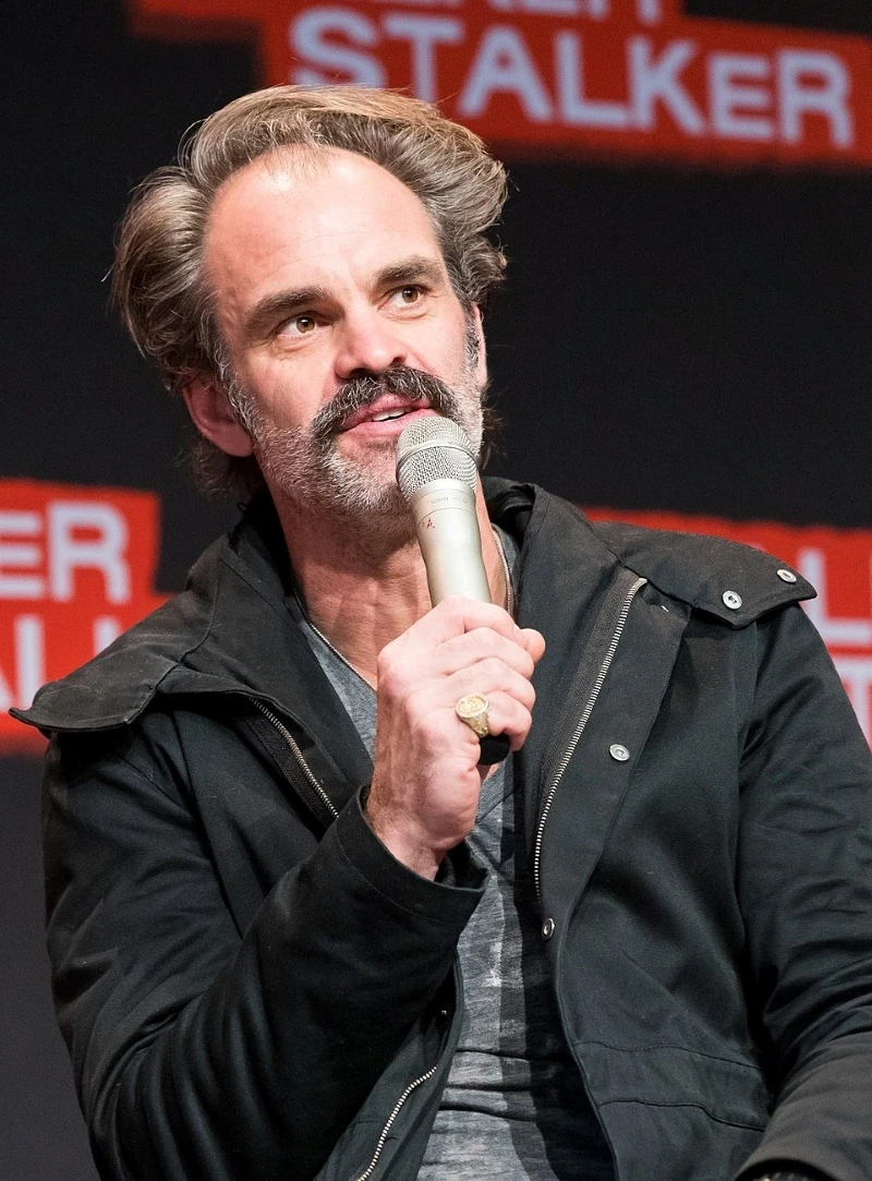 A person with a moustache, wearing a black jacket and grey shirt, speaks into a microphone against a black and red backdrop at a panel event, highlighting public speaking and engagement.