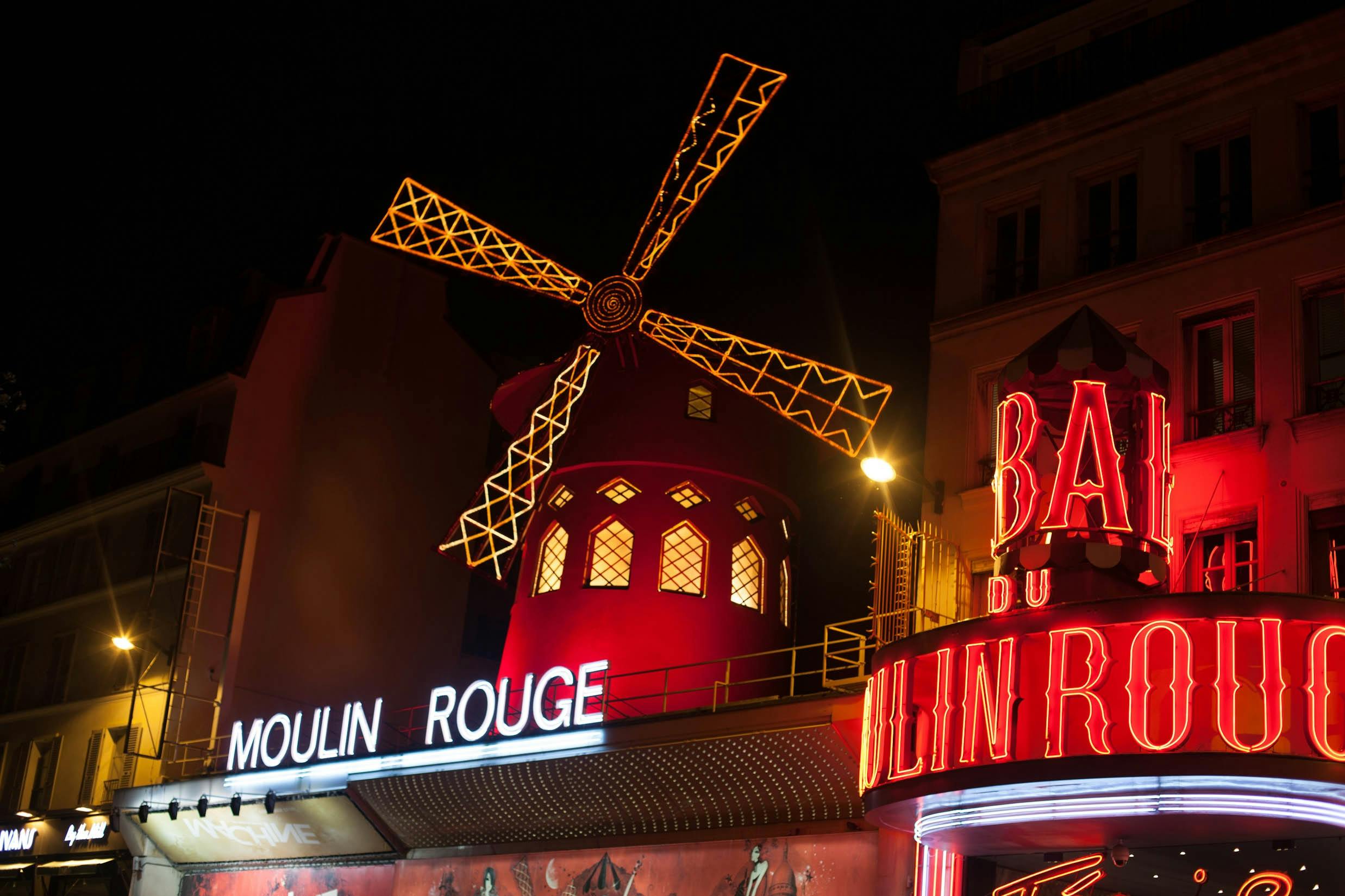 The illuminated red windmill of the Moulin Rouge cabaret in Paris glowing at night, surrounded by neon signs and vibrant lights.