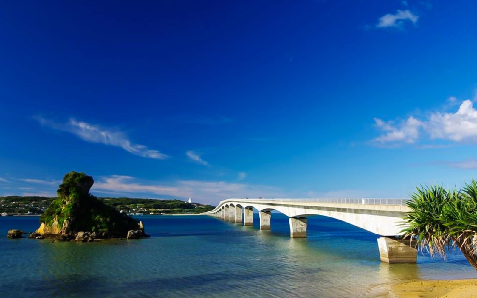 Bridge over clear blue water in Okinawa, Japan, with lush greenery and distant hills.