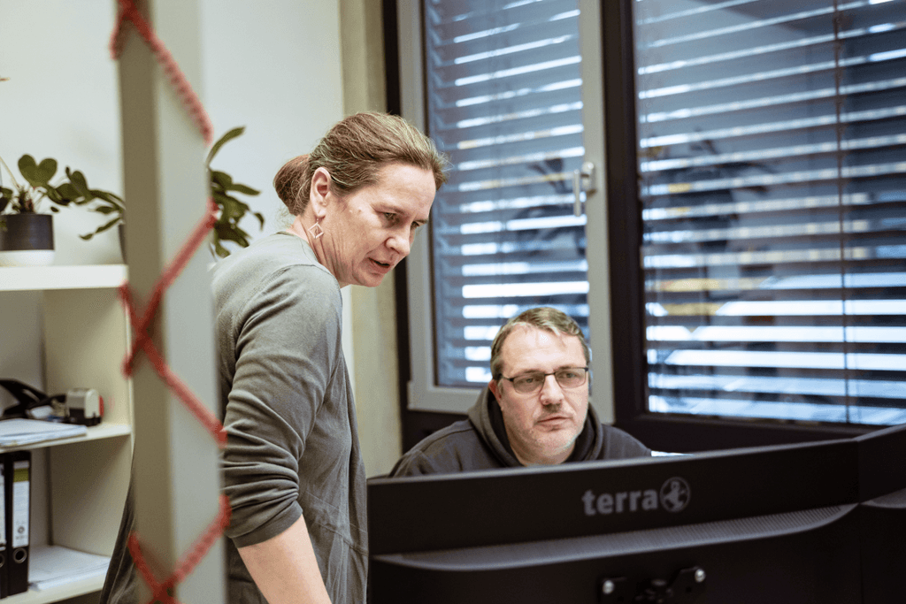 Zwei Mitarbeitende der kba Architekten und Ingenieure GmbH arbeiten gemeinsam am Computer; eine Frau beugt sich über den Bildschirm und erklärt etwas, während ihr Kollege konzentriert auf den Monitor schaut. Im Hintergrund stehen Regale und ein großes Fenster mit Jalousien.