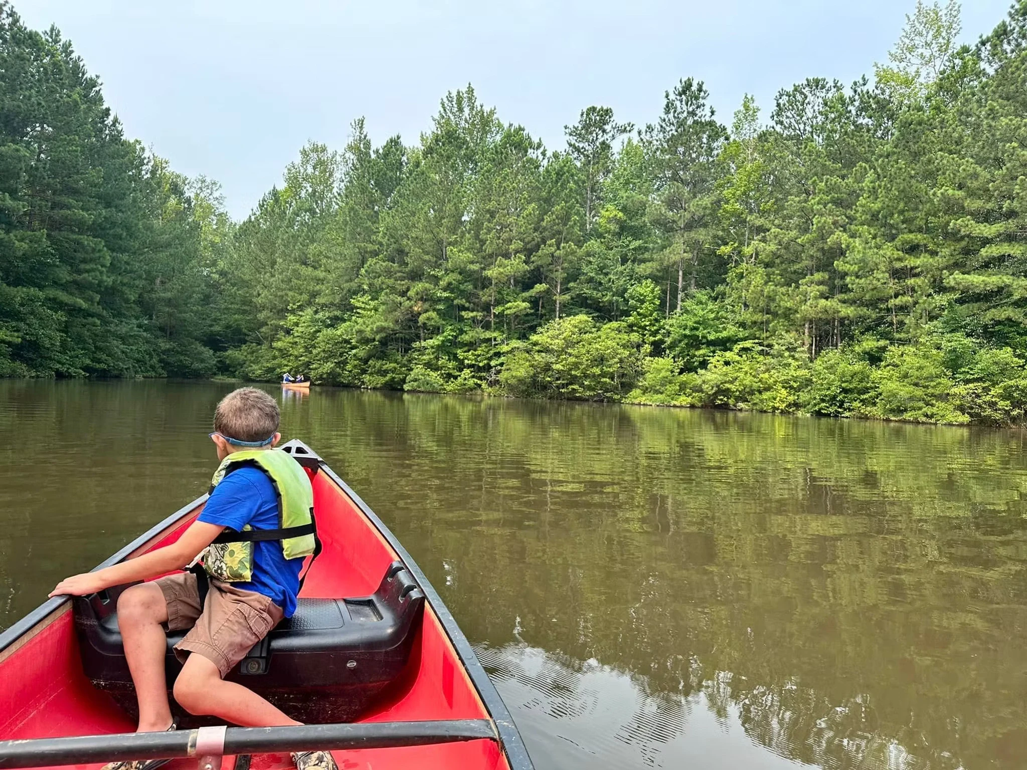 A boy sits in a red canoe paddling down a calm river surrounded by lush green trees.
