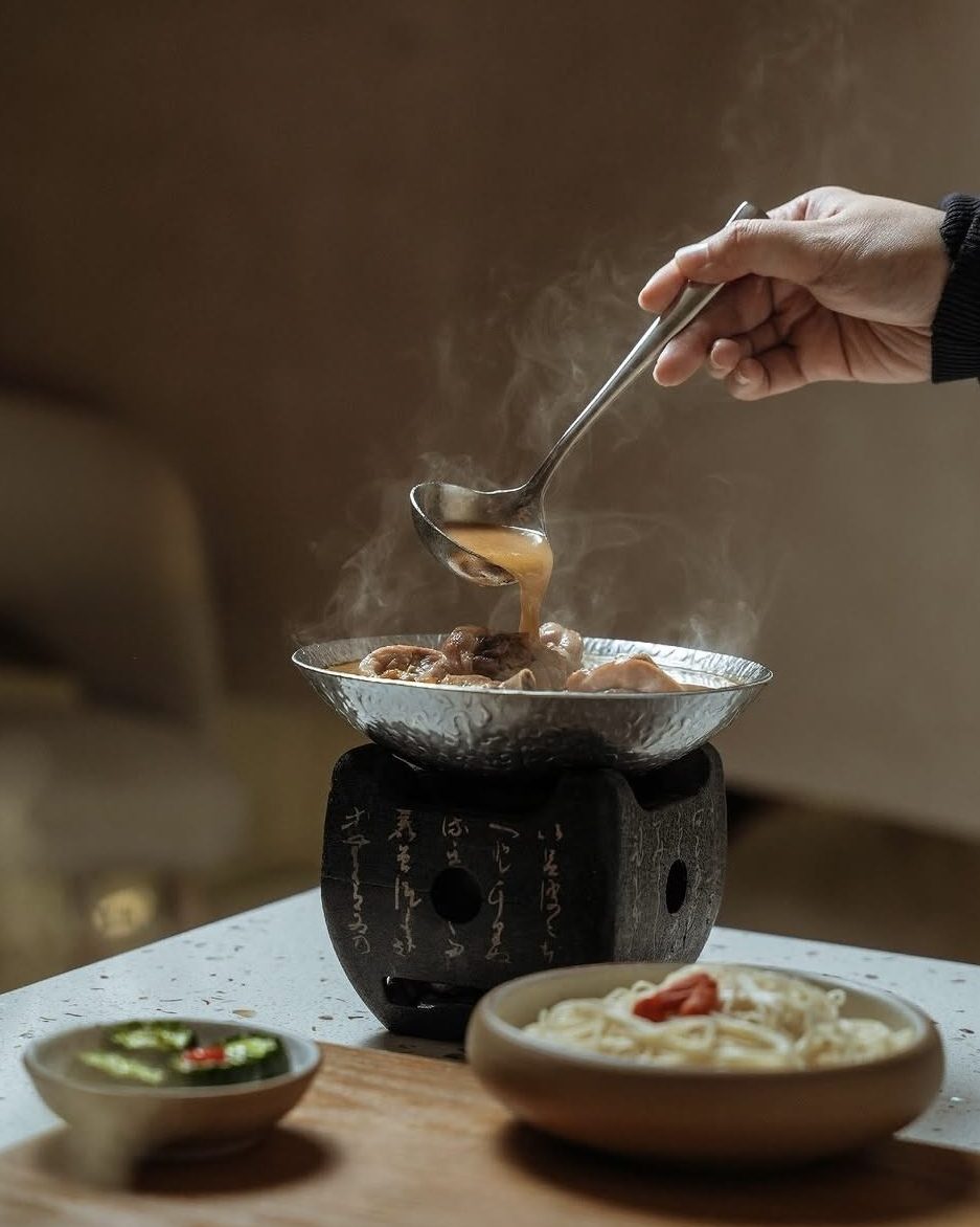 A hand holding a spoon serving steaming Three-Cup Chicken from a small pot, accompanied by bowls of noodles and sliced green chili on a wooden board.