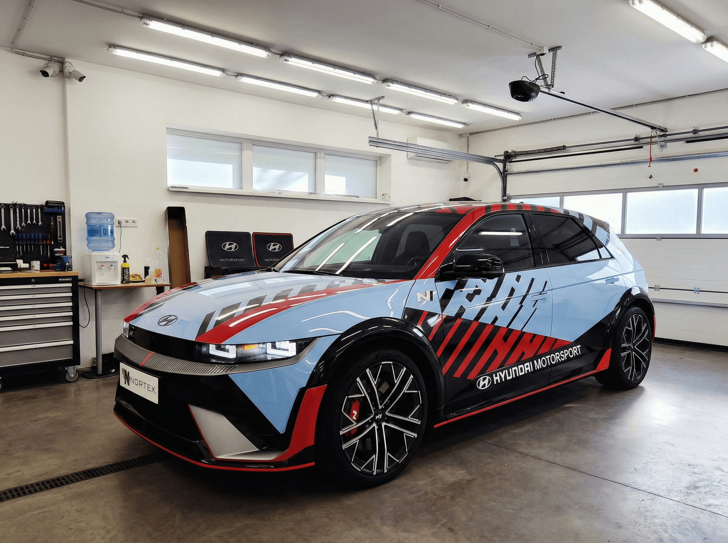 A NORTEK stúdióban egy Hyundai Ioniq 5 N nagy teljesítményű elektromos autó látható. Az autó bonyolult, egyedi racing livery dizájnt kapott kék, piros, fekete és fehér grafikai elemekkel. Az oldalajtókon egyedi verseny feliratok láthatók.