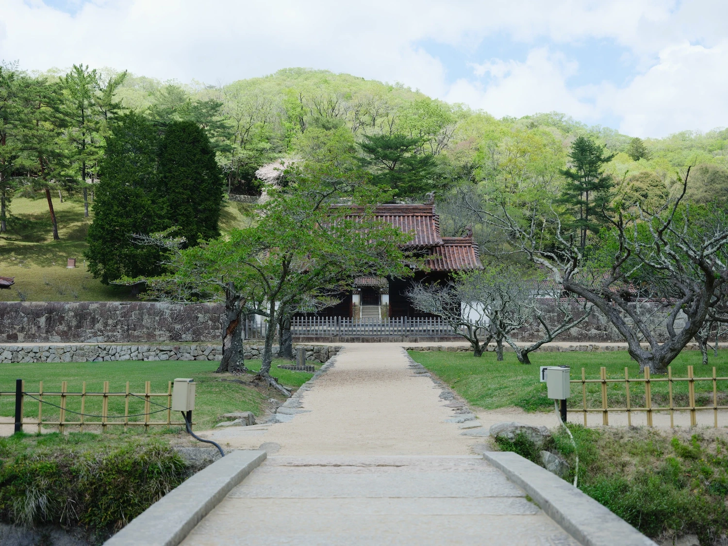 A view from the grounds of the former Shizutani School during Spring; there are trees lining the pathway and a hill in the back with more trees