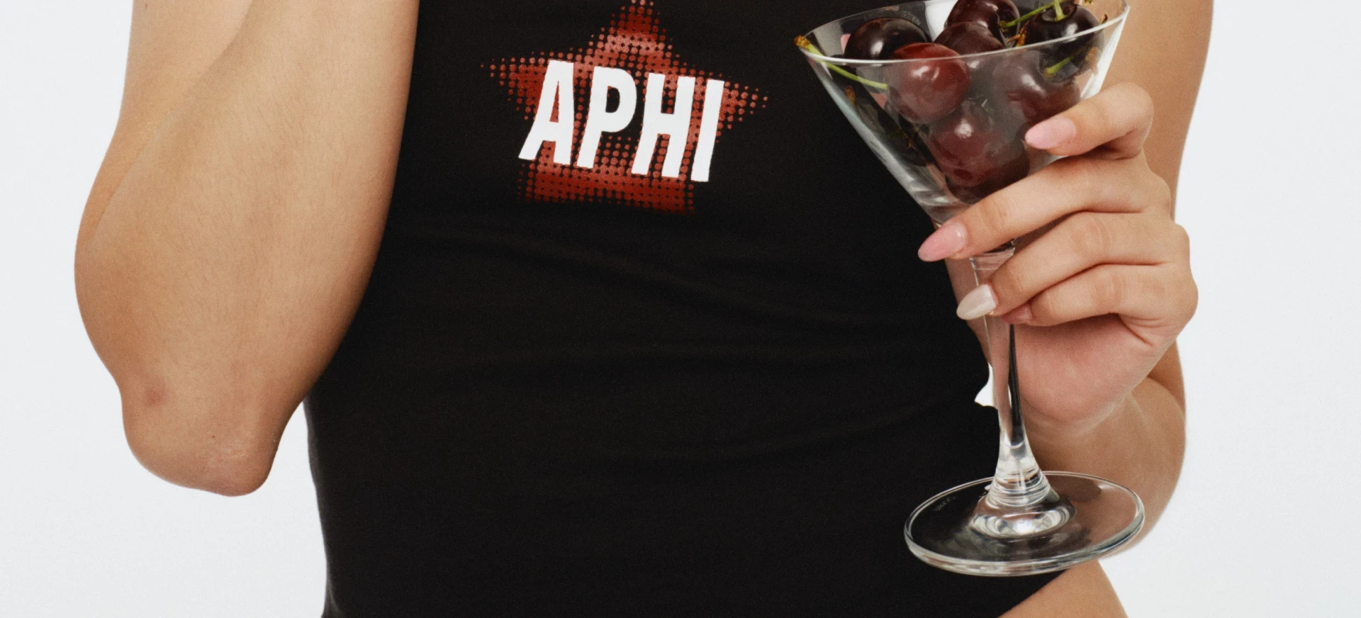 Close-up of a black tank top with “APHI” graphic, holding a martini glass filled with cherries