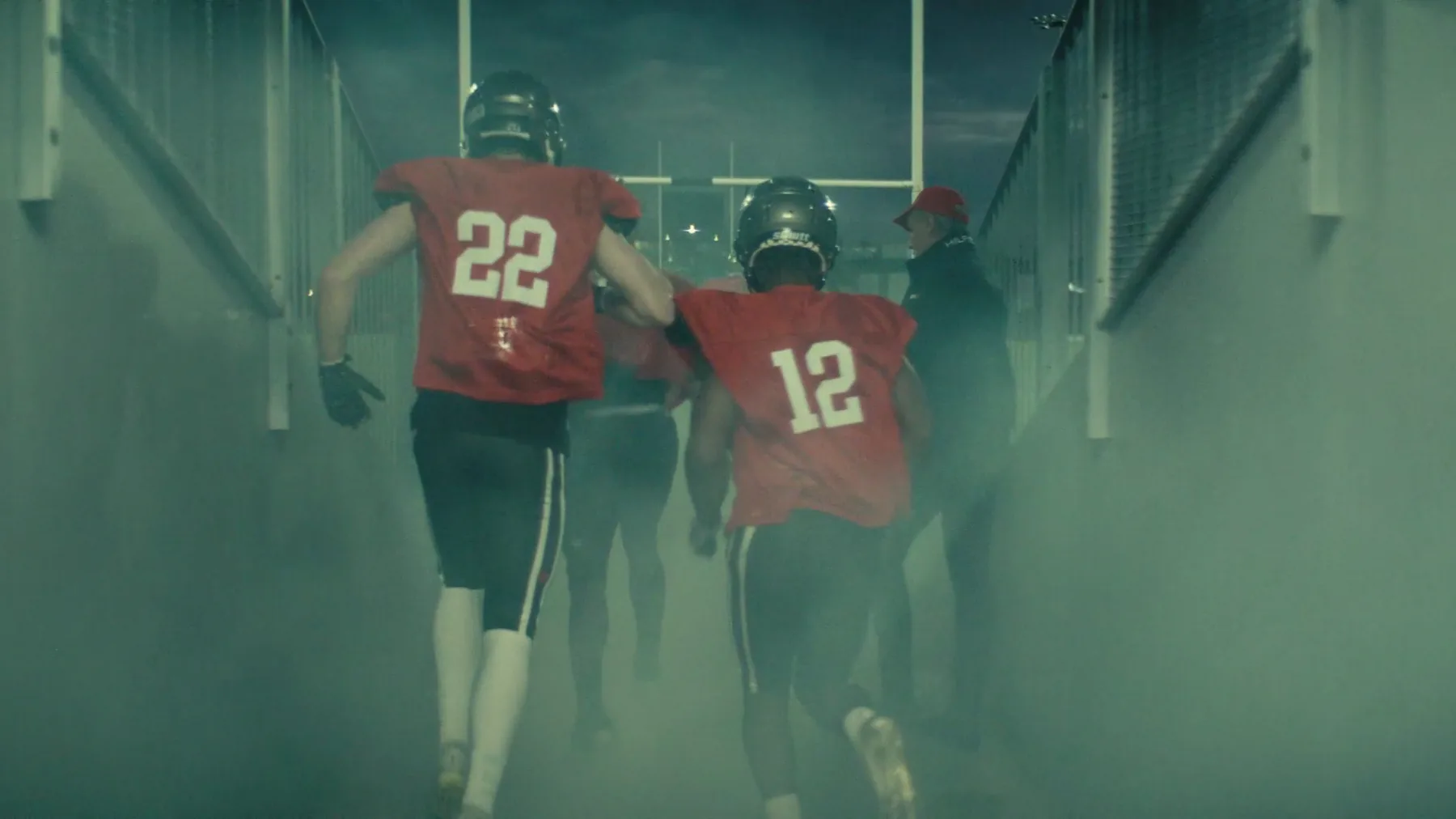 Two football players wearing red #22 and #12 jerseys run down a smoke-filled stadium tunnel toward the field, with a coach in a red cap standing to the side.