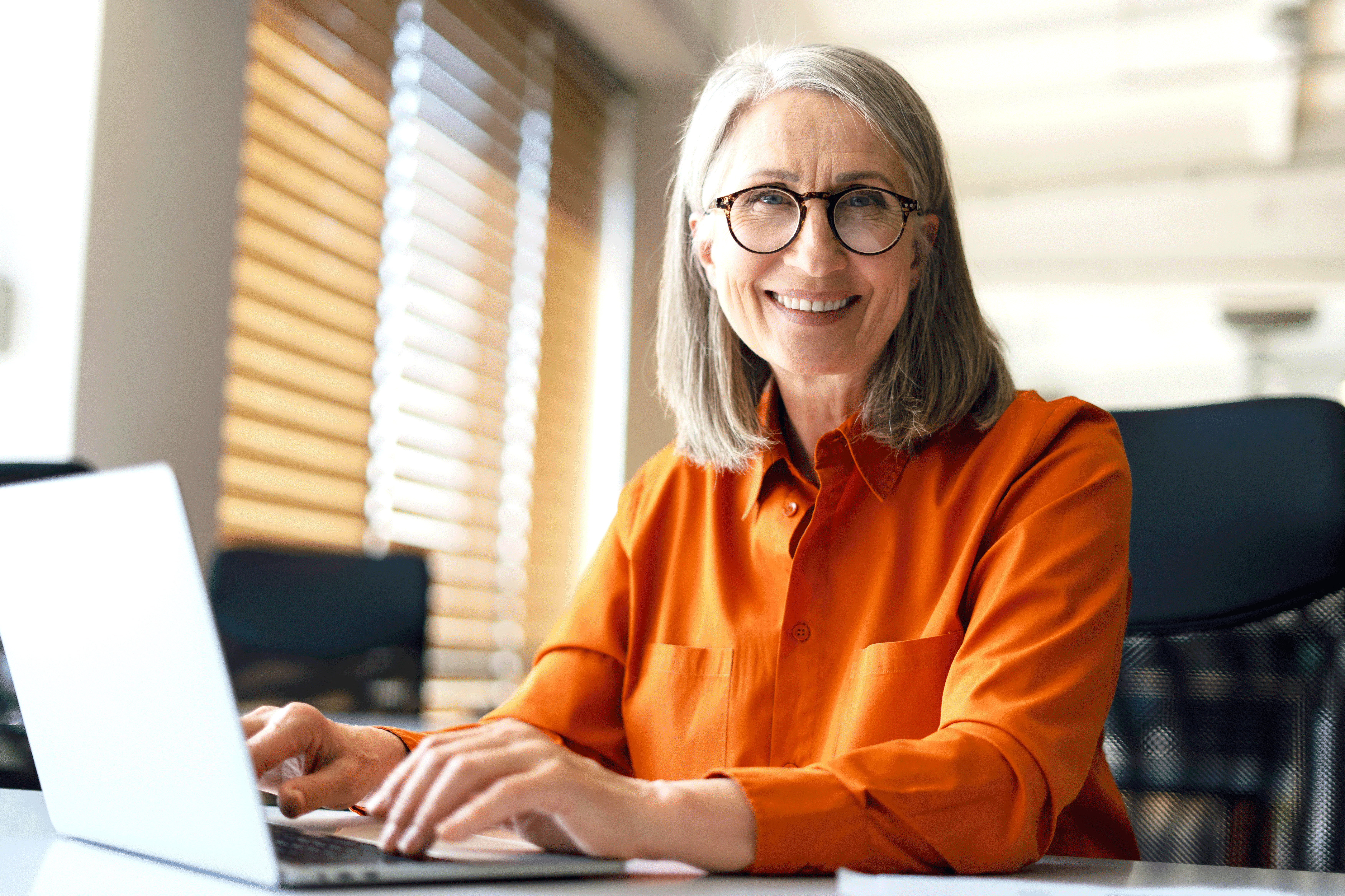 A smiling woman with gray hair and glasses wearing a bright orange button-down shirt sits at a desk while typing on a laptop in a sunlit office.