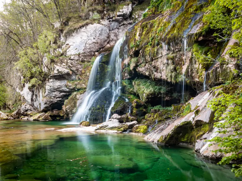 Virje waterfall in Soča river valley, Slovenia, flowing into a pool of emerald water through moss covered rocks.