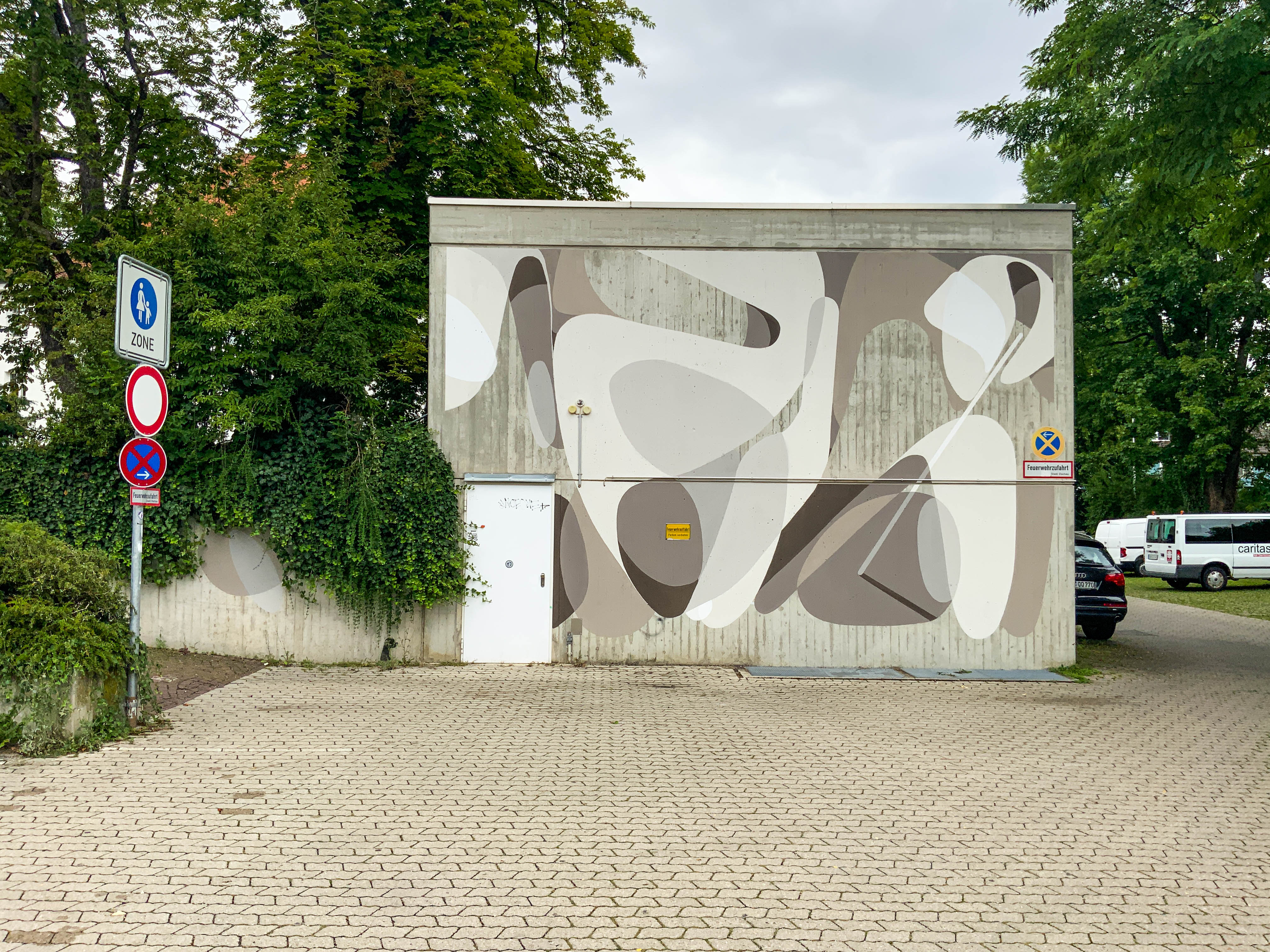 An abstract monochrome mural painted on a cube-shaped building, surrounded by green trees located on a parking area.
