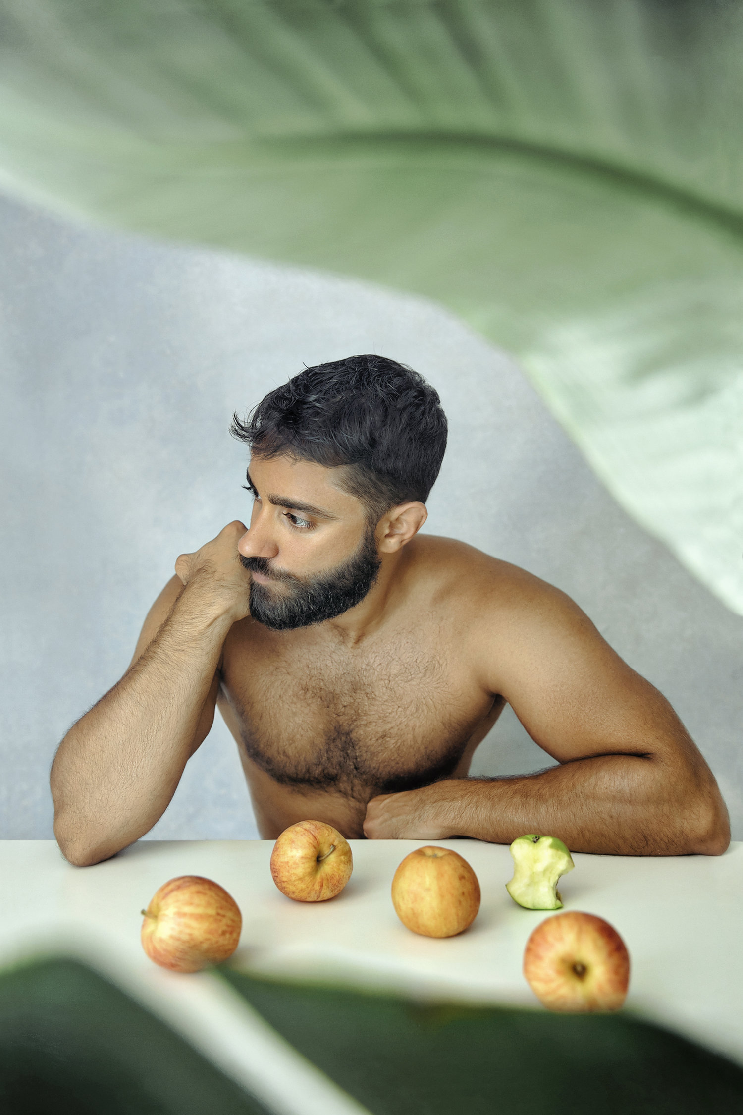 Julio Torres leaning on a table with apples, a contemplative pose amid green foliage, creating a serene, earthy atmosphere.