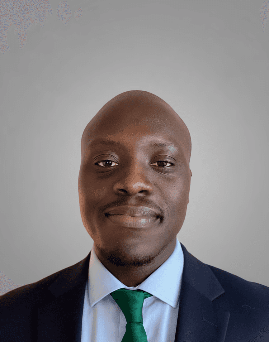 A smiling person with curly hair sits at a desk, looking at a laptop against a dark background.