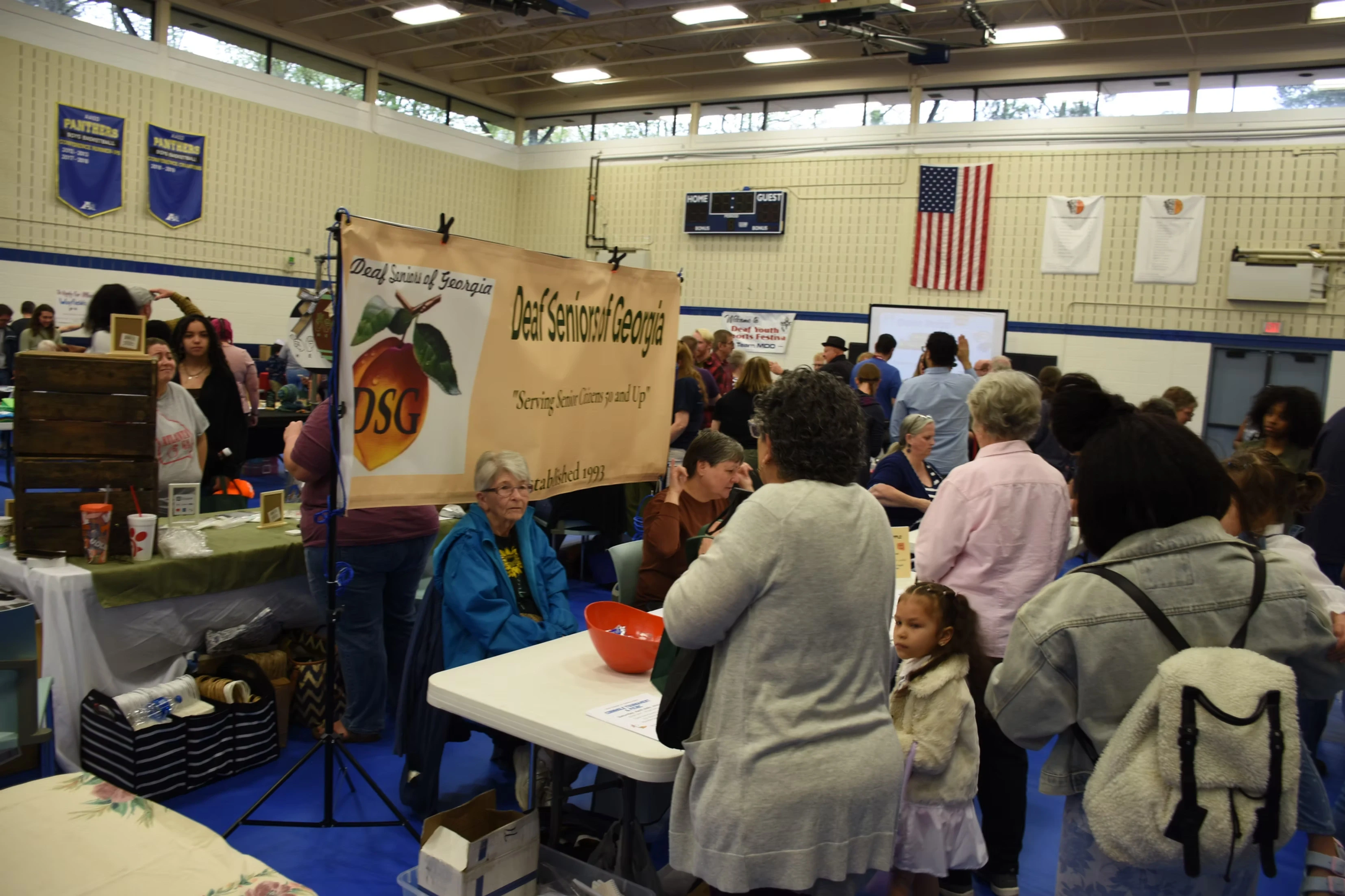 A bustling indoor market scene, with numerous people browsing various vendor tables. A banner for "Del Seymour Georgia" is prominently displayed.